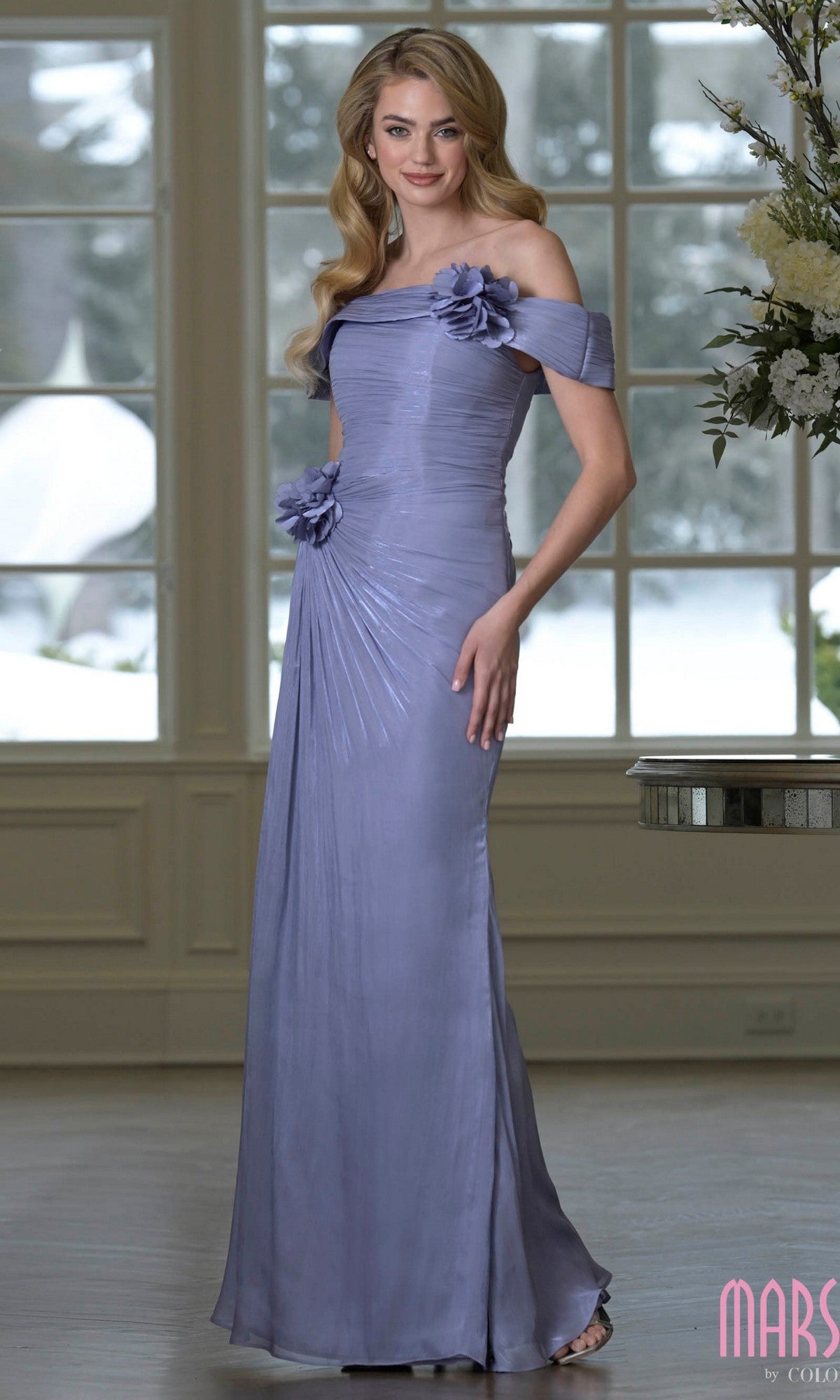 A woman stands indoors wearing the Marsoni M353—an elegant, floor-length, off-the-shoulder dusty blue dress with 3D floral accents and pleated detailing. Snowy scenery is visible through the windows behind her.